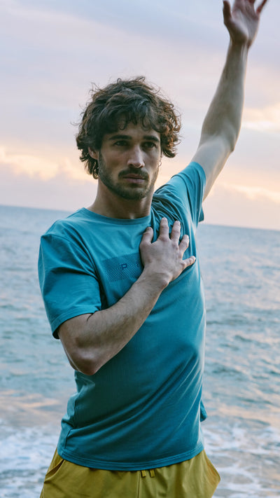 Hombre realizando un estiramiento con el brazo extendido, vistiendo una camiseta azul y un bañador amarillo de picsil









